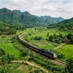 Passenger train winding through green rice fields beneath forested mountains
